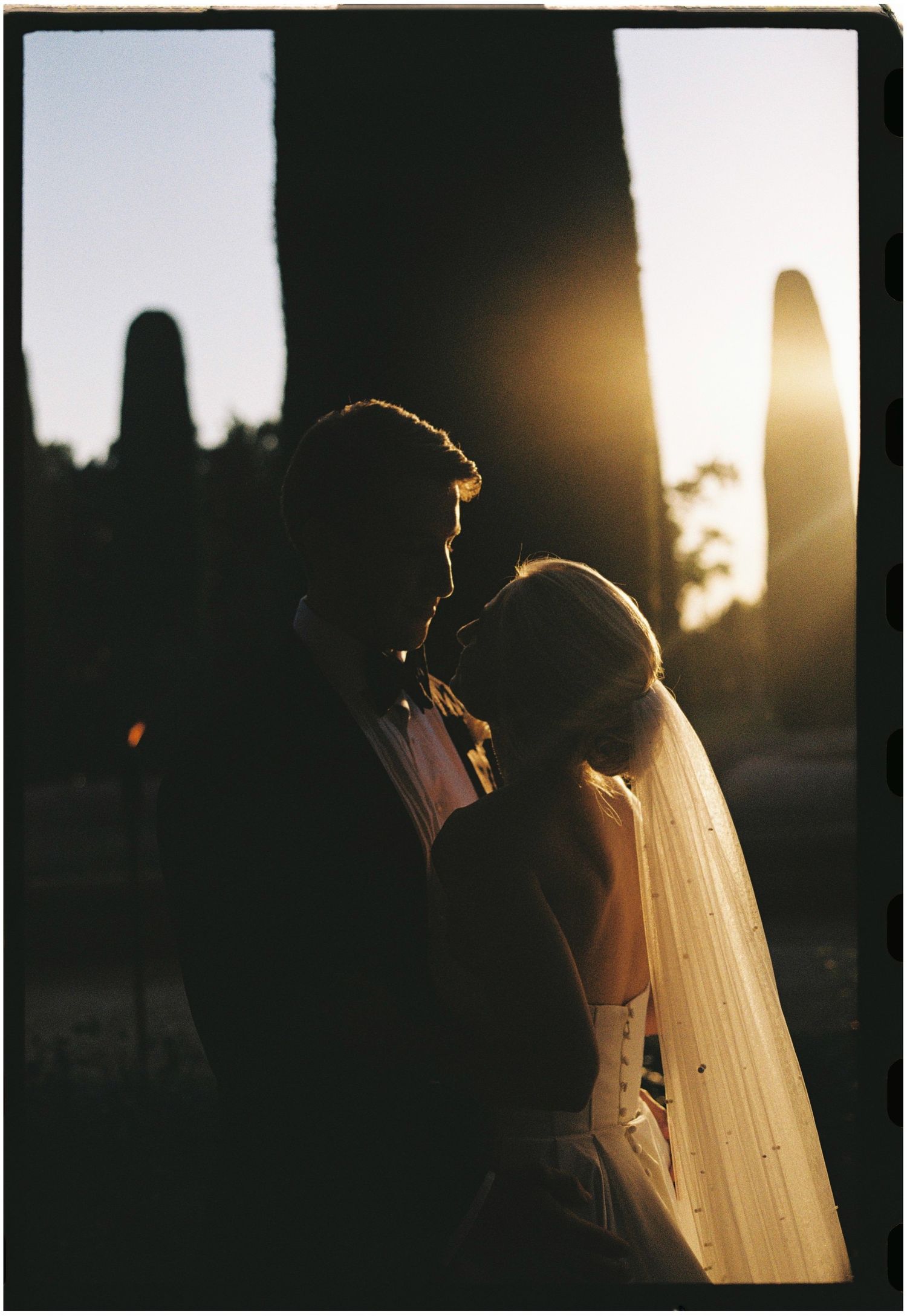 Silhouetted couple embracing against golden sunset with veil flowing in dramatic backlight.