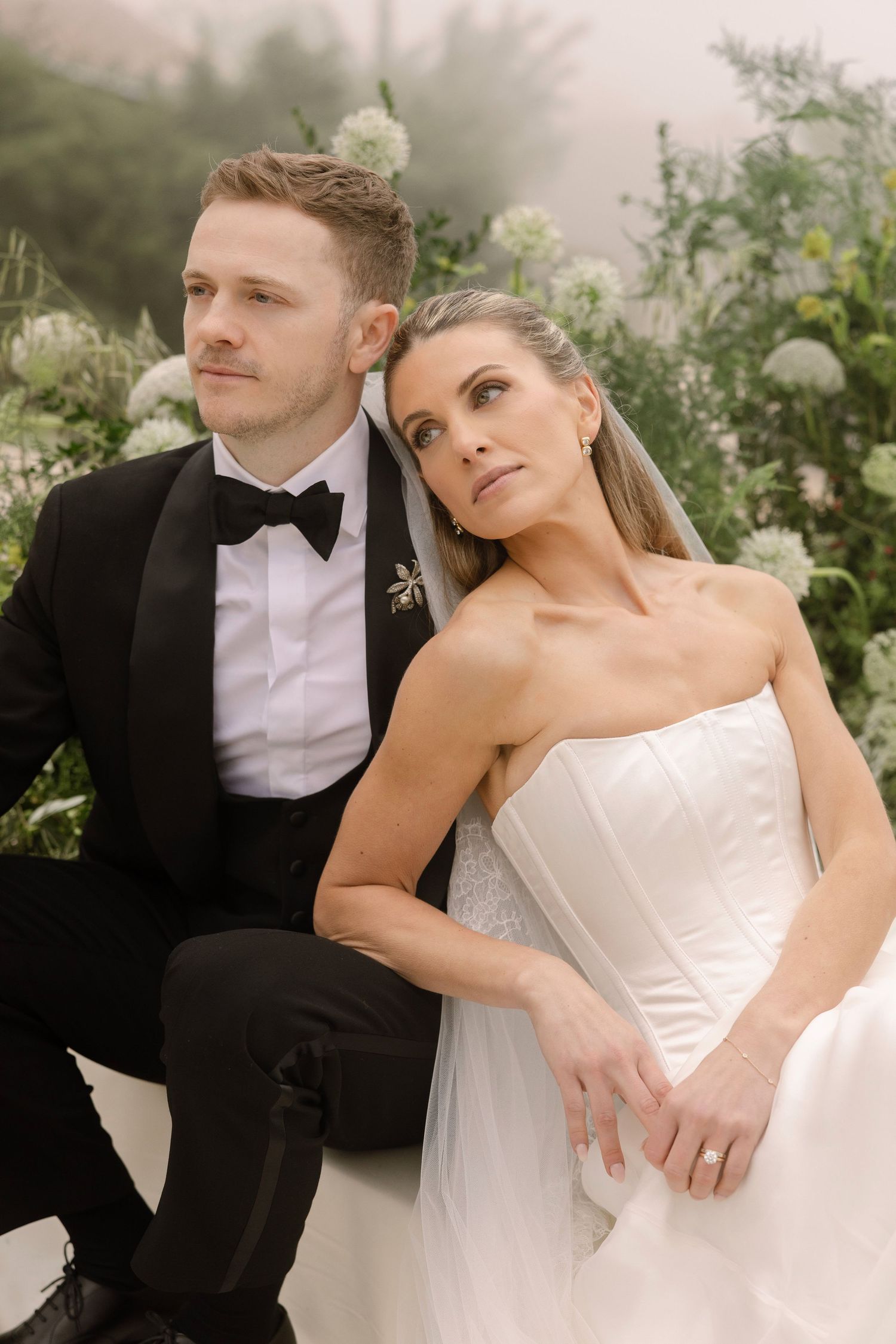 A bride and groom pose elegantly outdoors, she in a strapless white gown with veil, he in a classic black tuxedo.