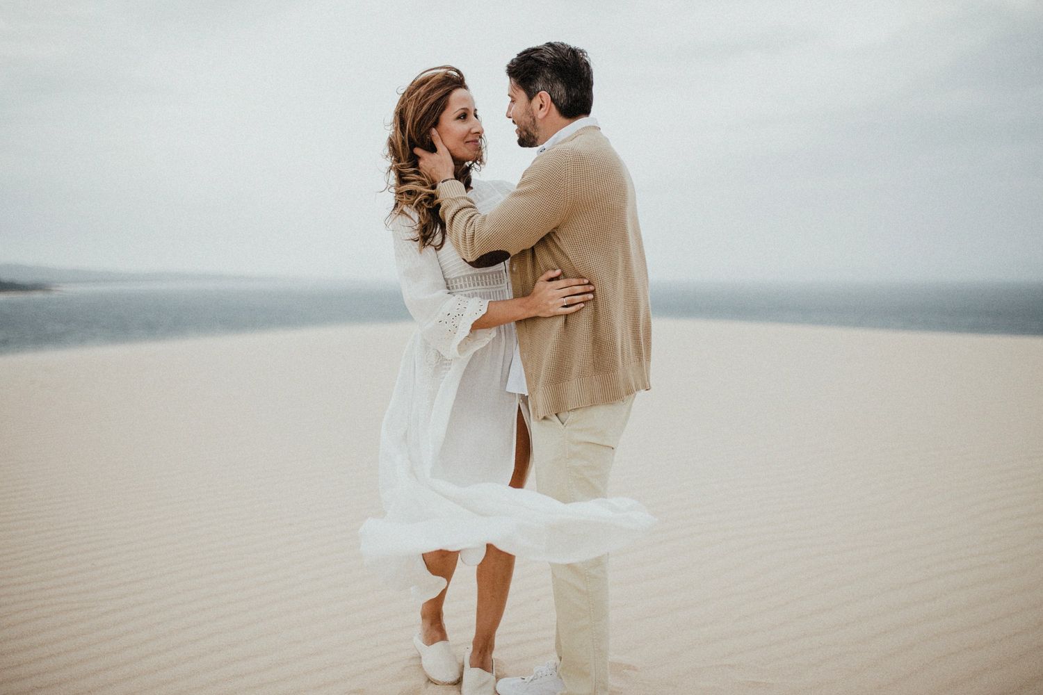 Couple dancing together on sand dunes with flowing white dress in motion.