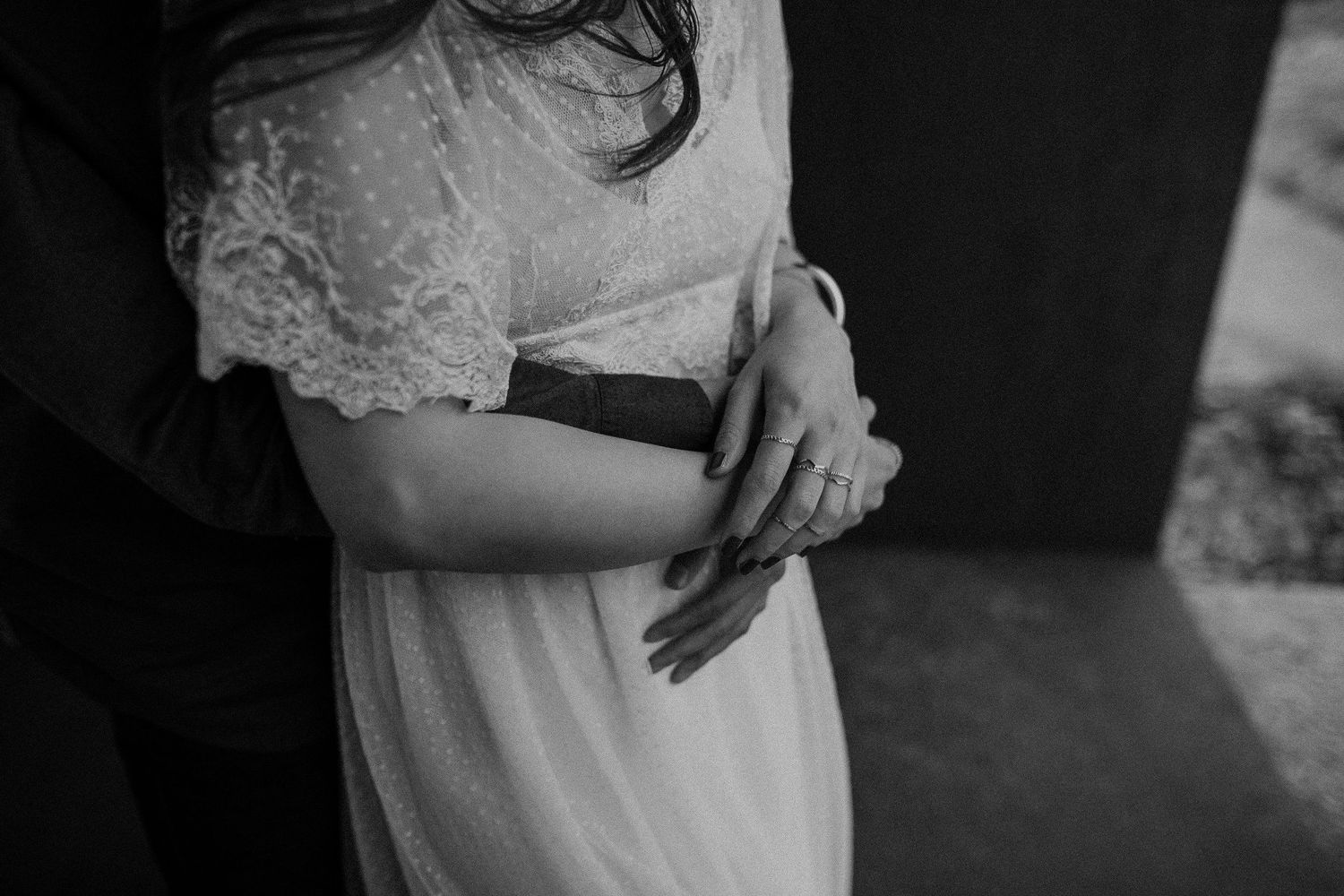 A black and white close-up of hands clasped together while wearing elegant wedding attire.