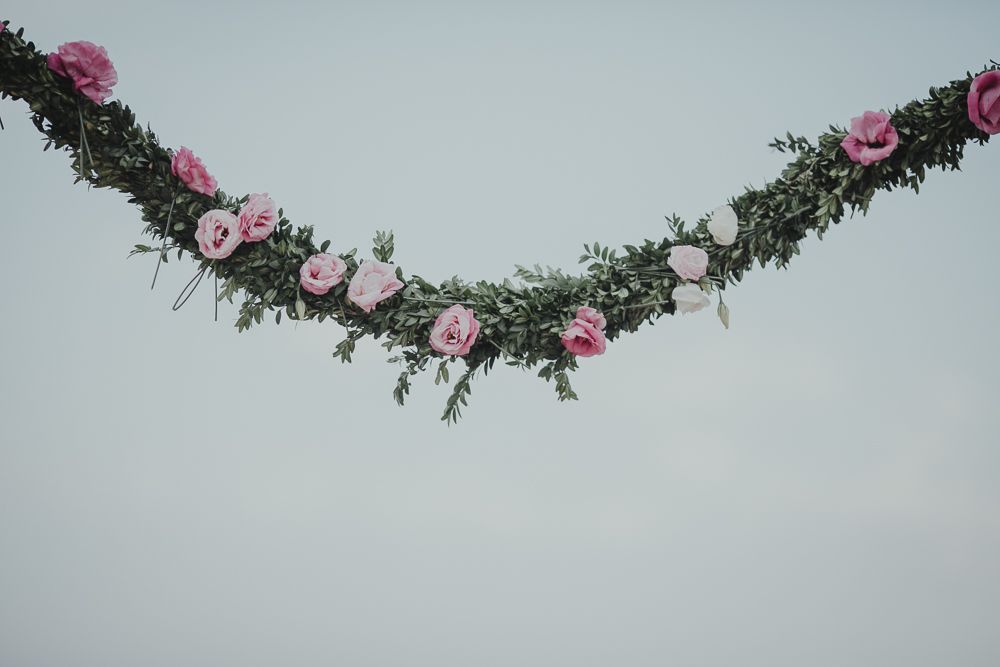 Delicate floral garland with pink roses and greenery hanging against a pale grey sky.