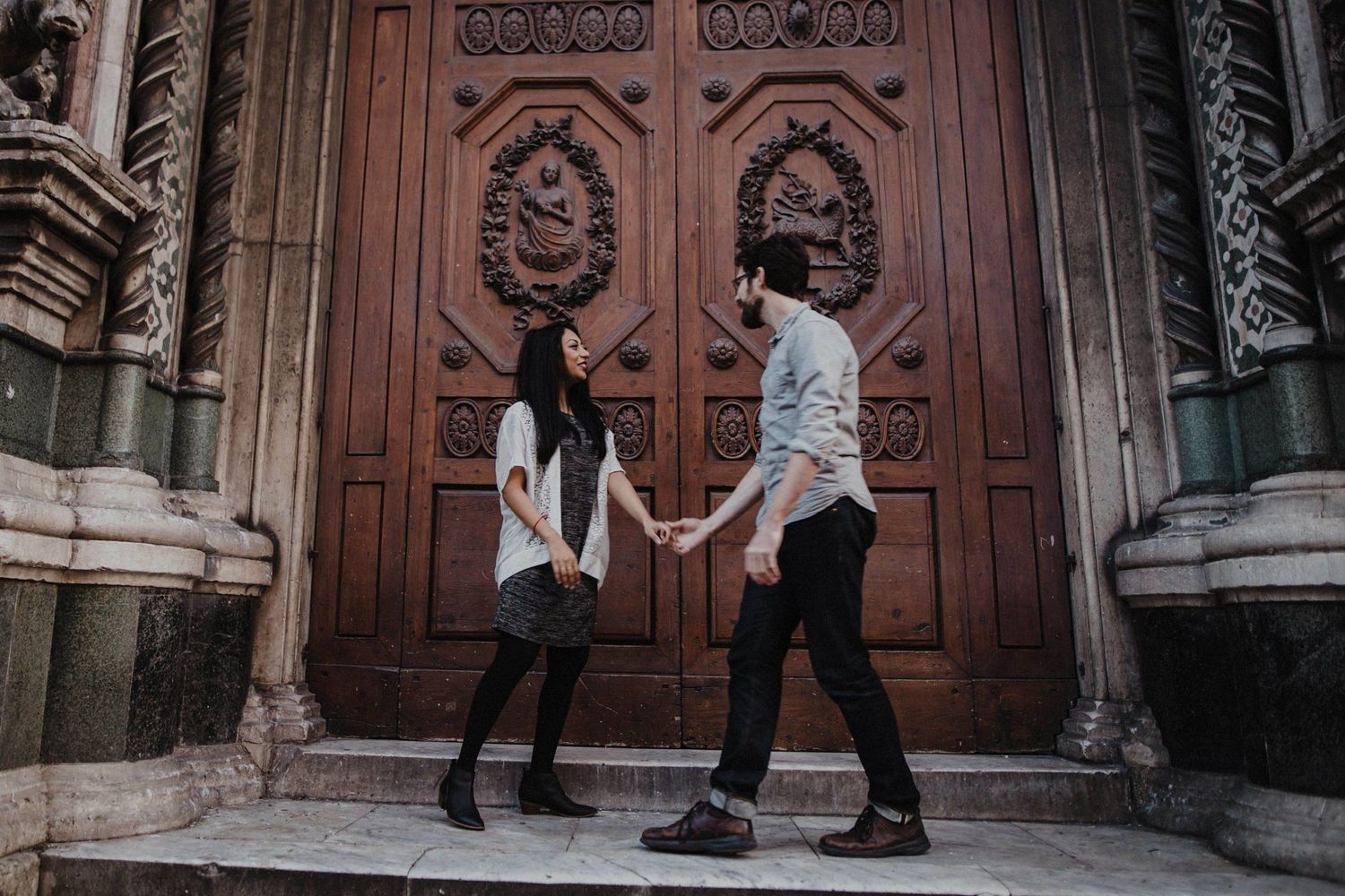 Couple standing on church steps holding hands against large ornate wooden doors.