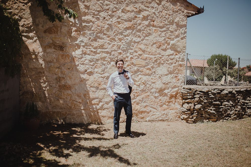 Someone in formal attire stands against a textured stone wall outside on a sunny day.