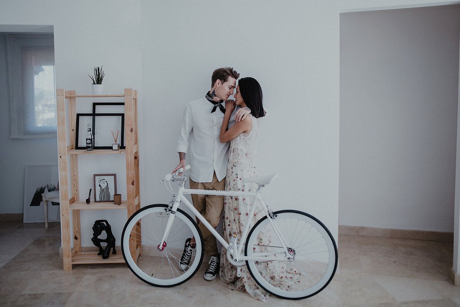A romantic couple embracing next to a white bicycle and wooden shelf unit.