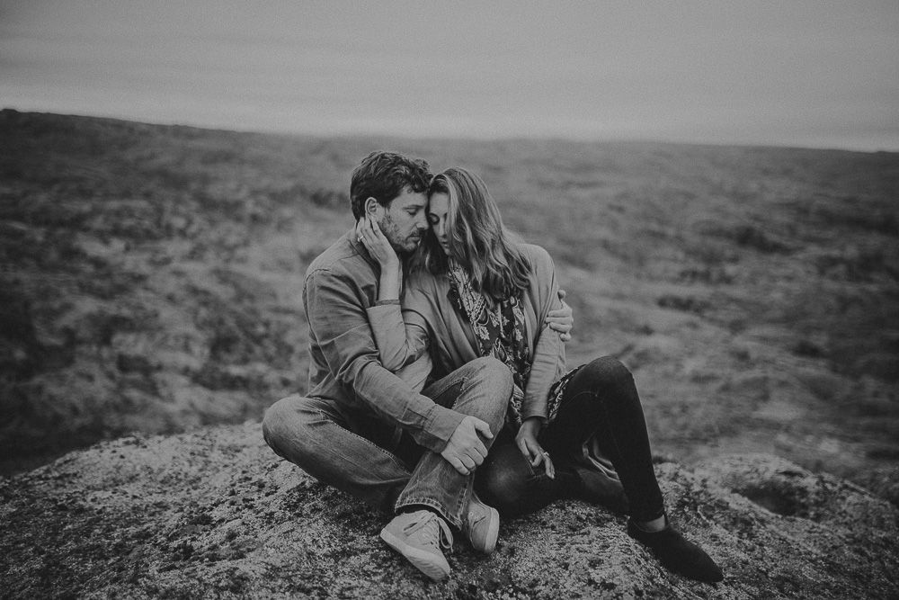 Intimate couple embraces while sitting on rugged cliffside during moody overcast day.