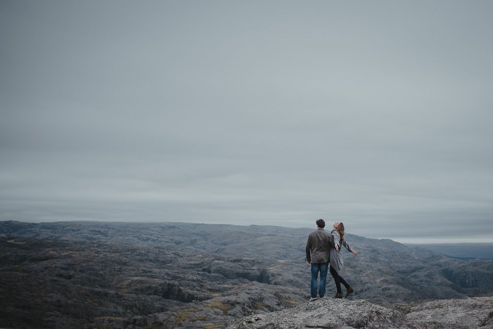 A couple stands together on a rocky mountain overlook with vast hills stretching to the horizon.