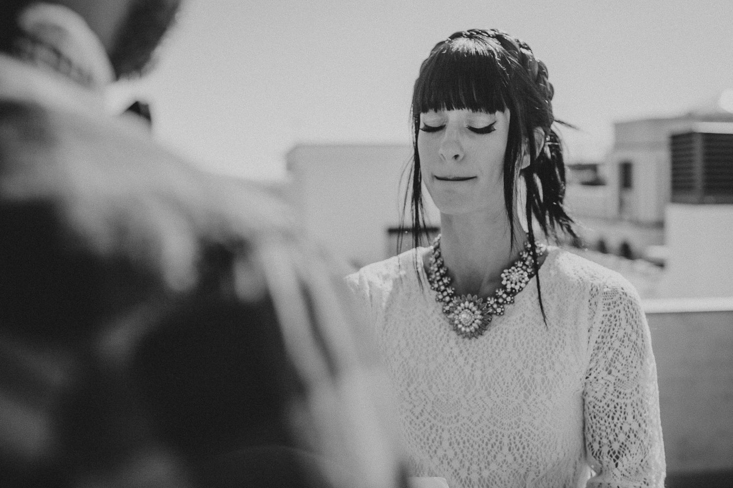A woman in white lace and statement jewelry stands against an urban rooftop backdrop.
