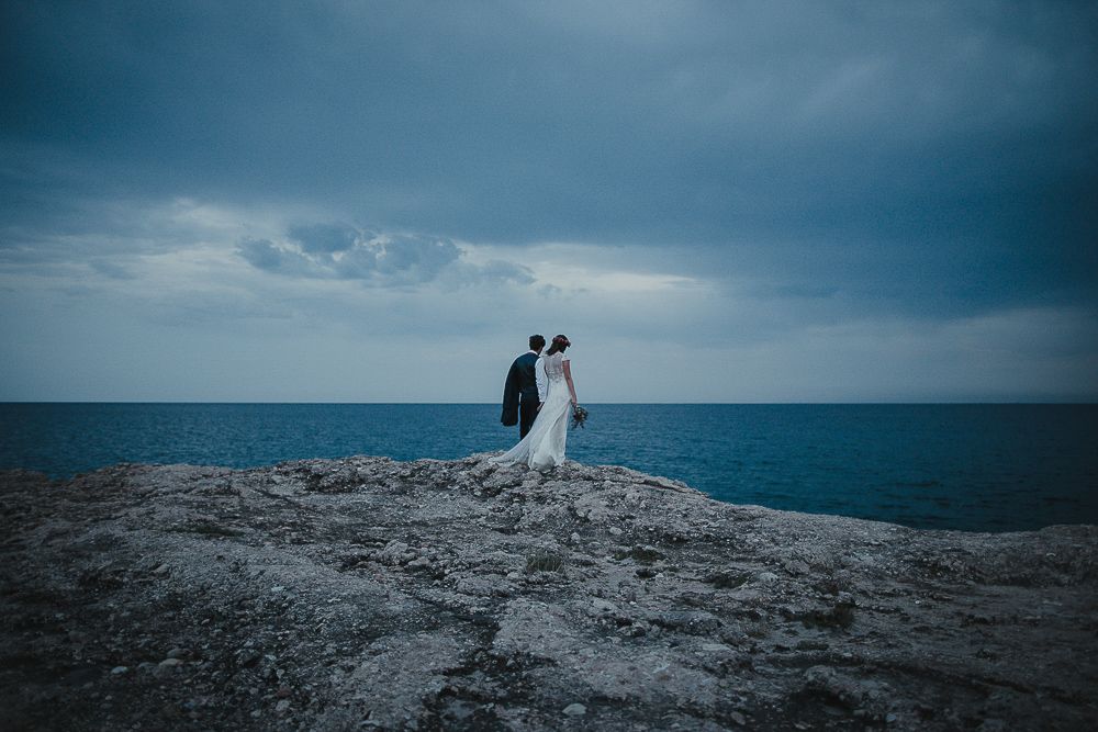 Silhouettes of a wedding couple standing on a rocky cliff against a dramatic ocean backdrop.