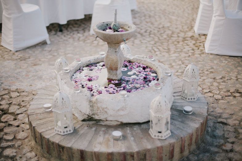 A decorative stone fountain filled with flower petals and surrounded by glass containers on a cobblestone surface.