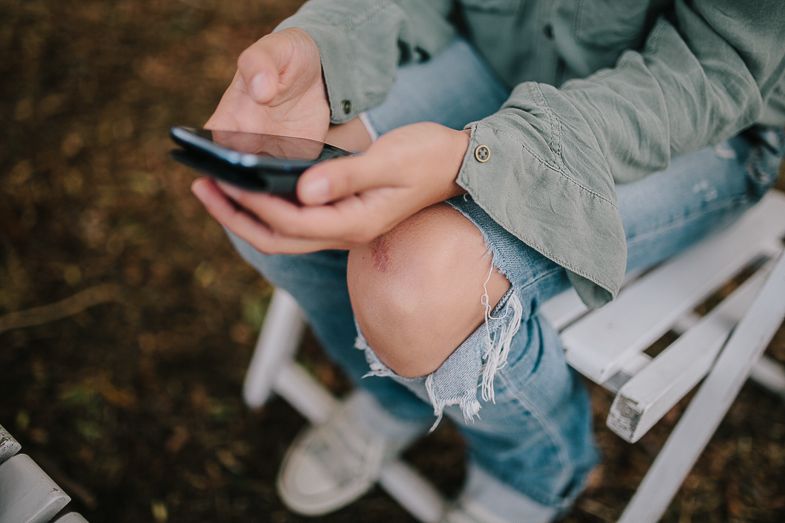 Close-up view of hands holding a mobile phone while seated on a white folding chair outdoors.