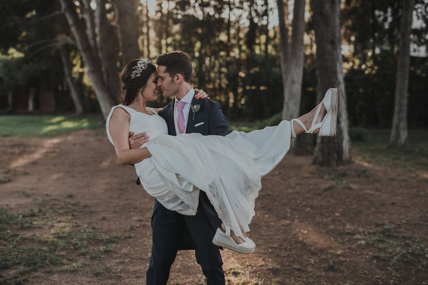 A groom lifts his bride in a playful pose on a tree-lined path during sunset.