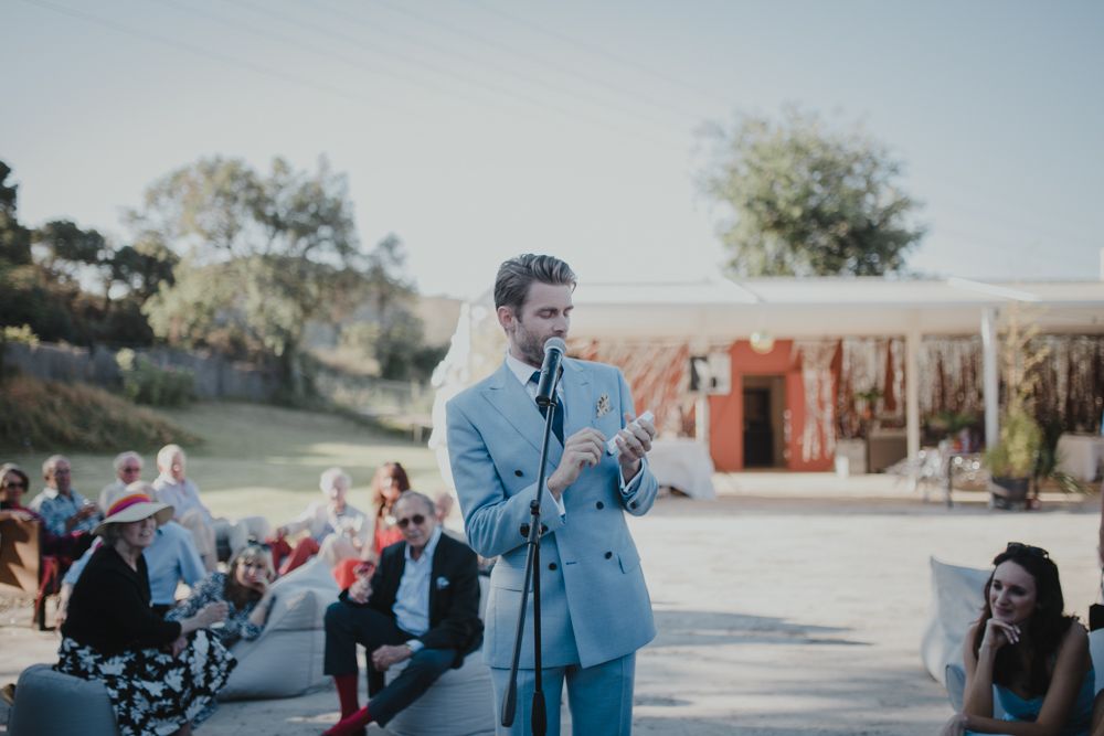 Someone in a light blue suit speaks into a microphone at an outdoor event.