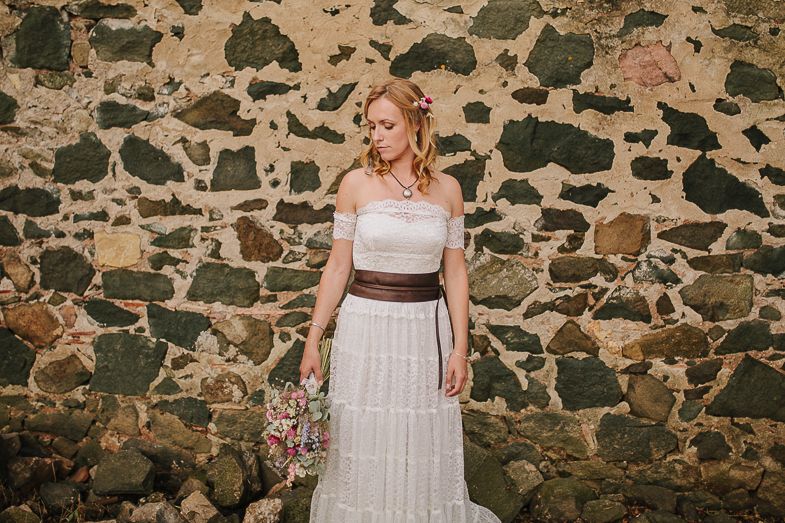 Bride in a white dress with brown belt poses against rustic stone wall while holding bouquet.