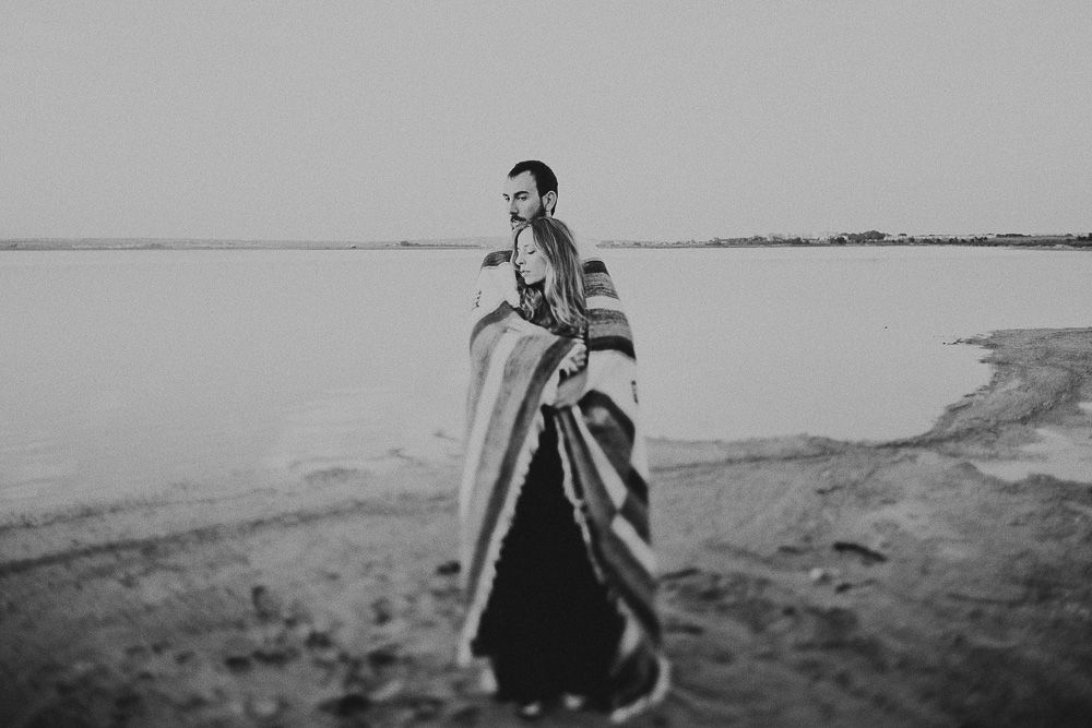 Person wrapped in blanket stands on shoreline in dramatic black and white photo.