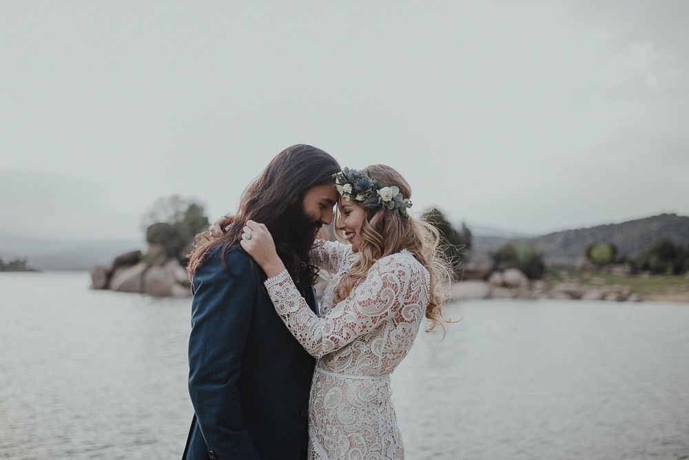 Wedding couple shares tender moment on rocky lakeshore under overcast sky.