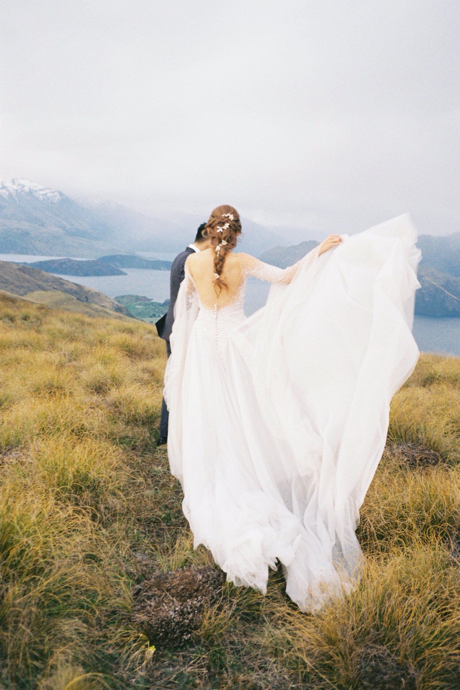 Flowing white wedding dress billows in wind on mountain peak with lake view.