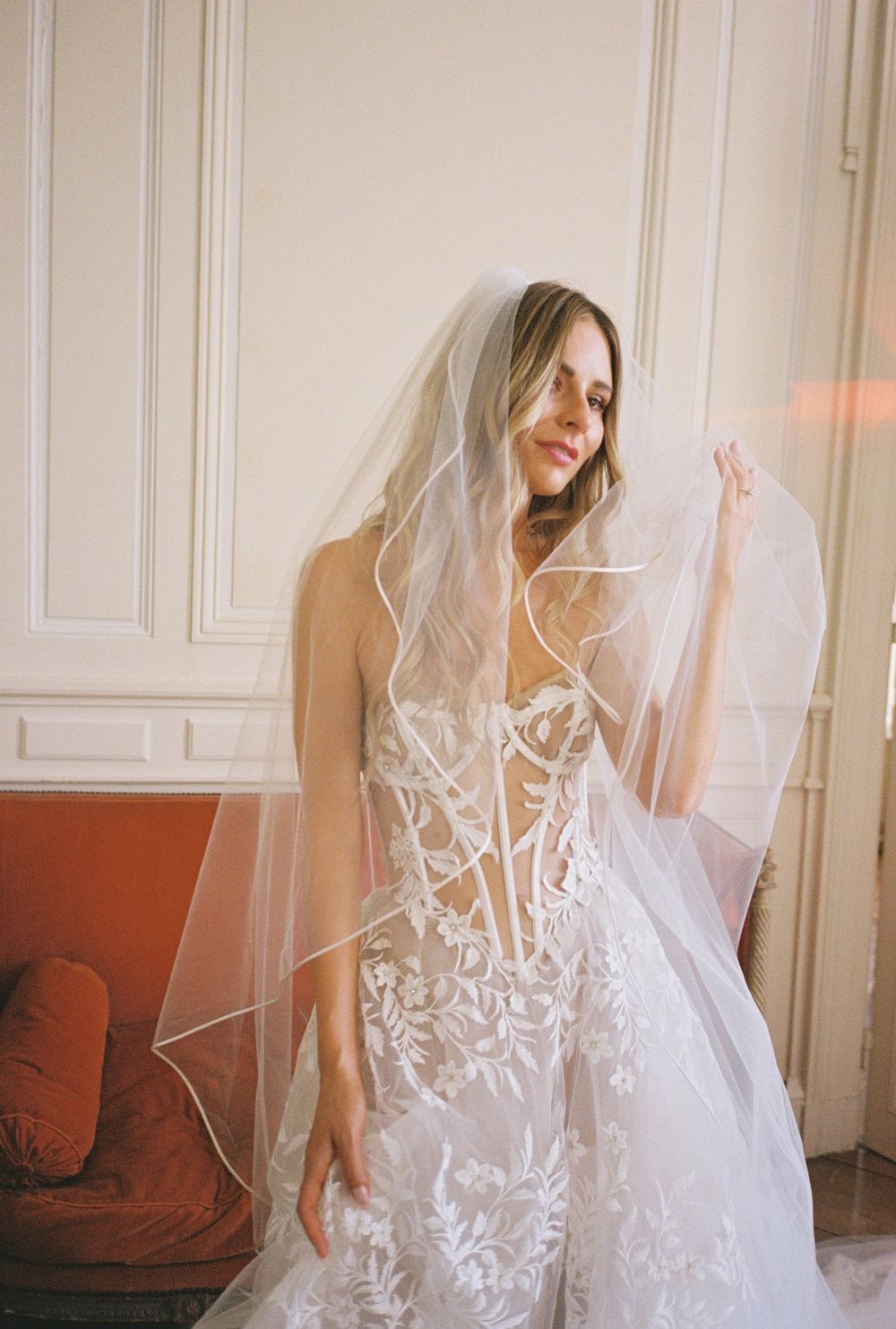 Ethereal bridal portrait featuring flowing lace wedding gown against white wall.