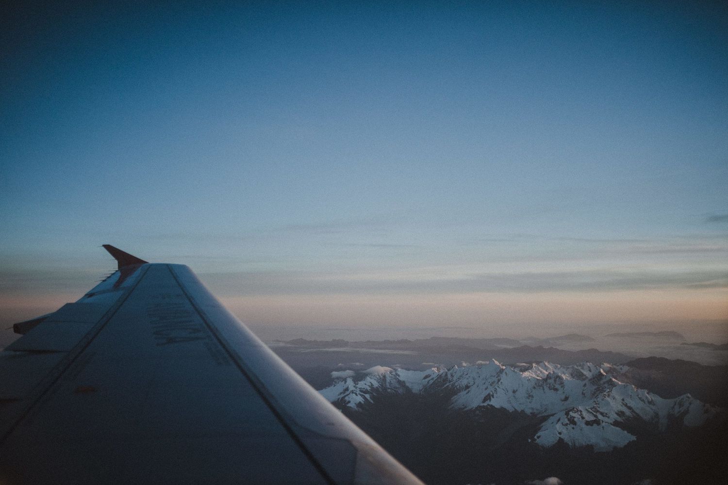 Aircraft wing silhouetted against blue sky with snow-capped mountains visible below.