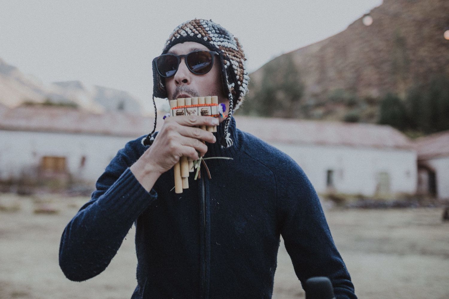 A person in winter clothing and sunglasses drinks from a glass outdoors.