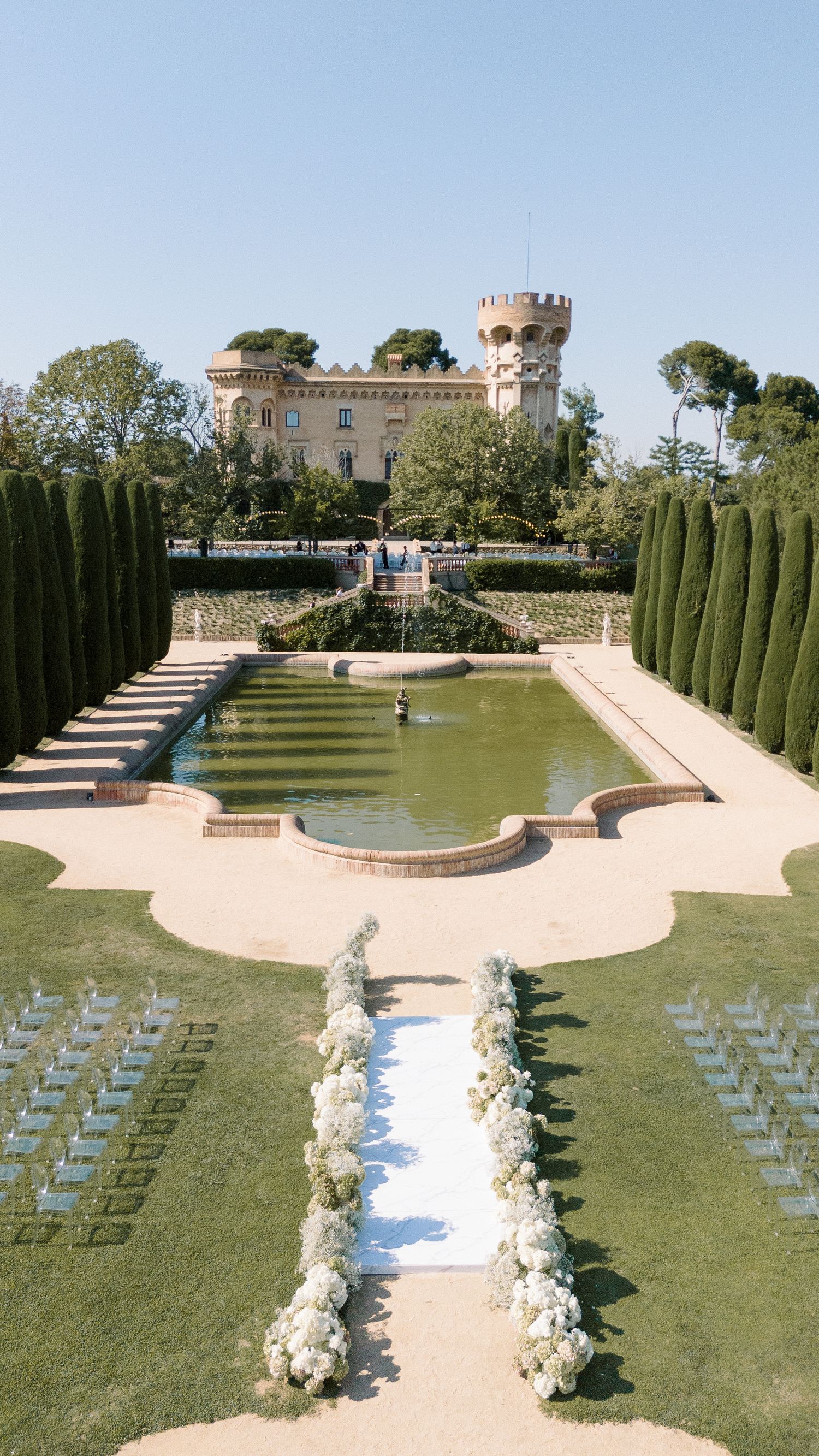 Castle-like venue with manicured gardens, cypress trees, and reflecting pool surrounded by ceremony seating.