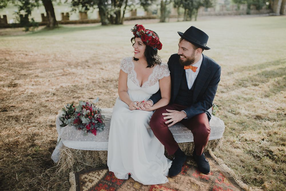 A couple shares a moment while sitting on a rustic bench with a purple floral bouquet.