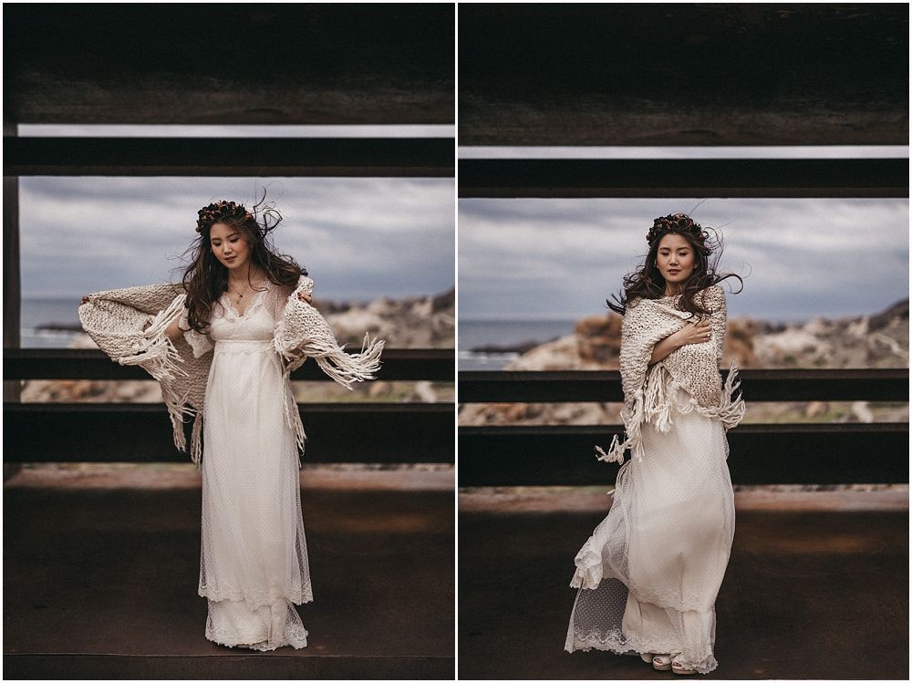A bride in a flowing white dress poses dramatically in a concrete viewing area with ocean backdrop.