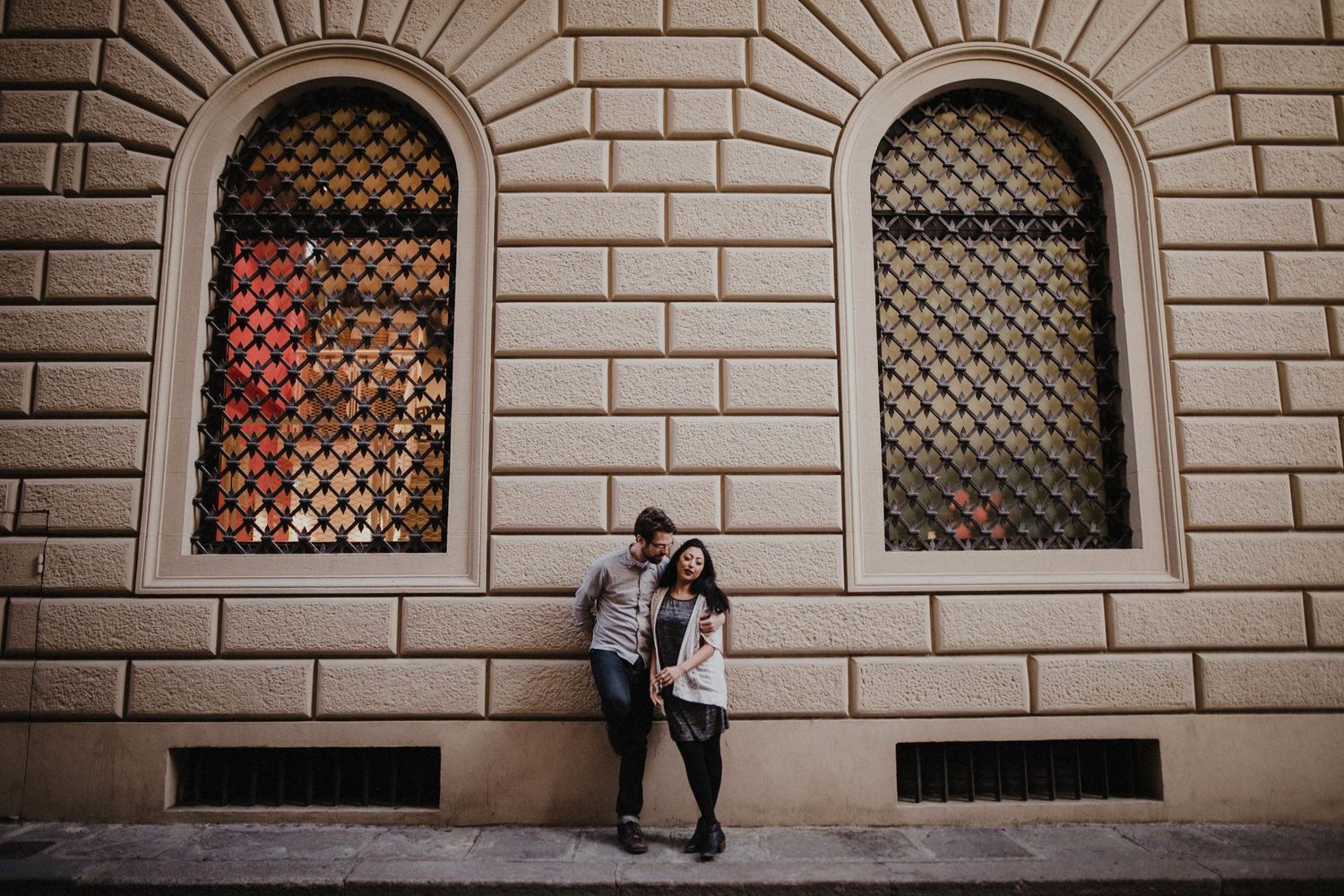 A couple stands beneath ornate arched windows on a historic building facade.