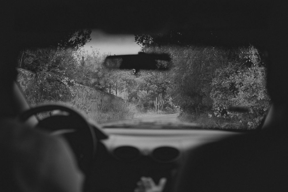 Black and white view through a car windshield showing a rainy rural road ahead.