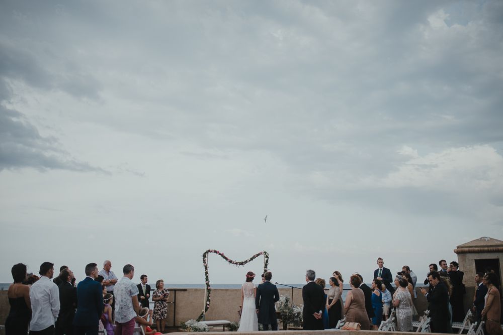 Wedding guests gather for an outdoor ceremony against a cloudy sky on a rooftop terrace.