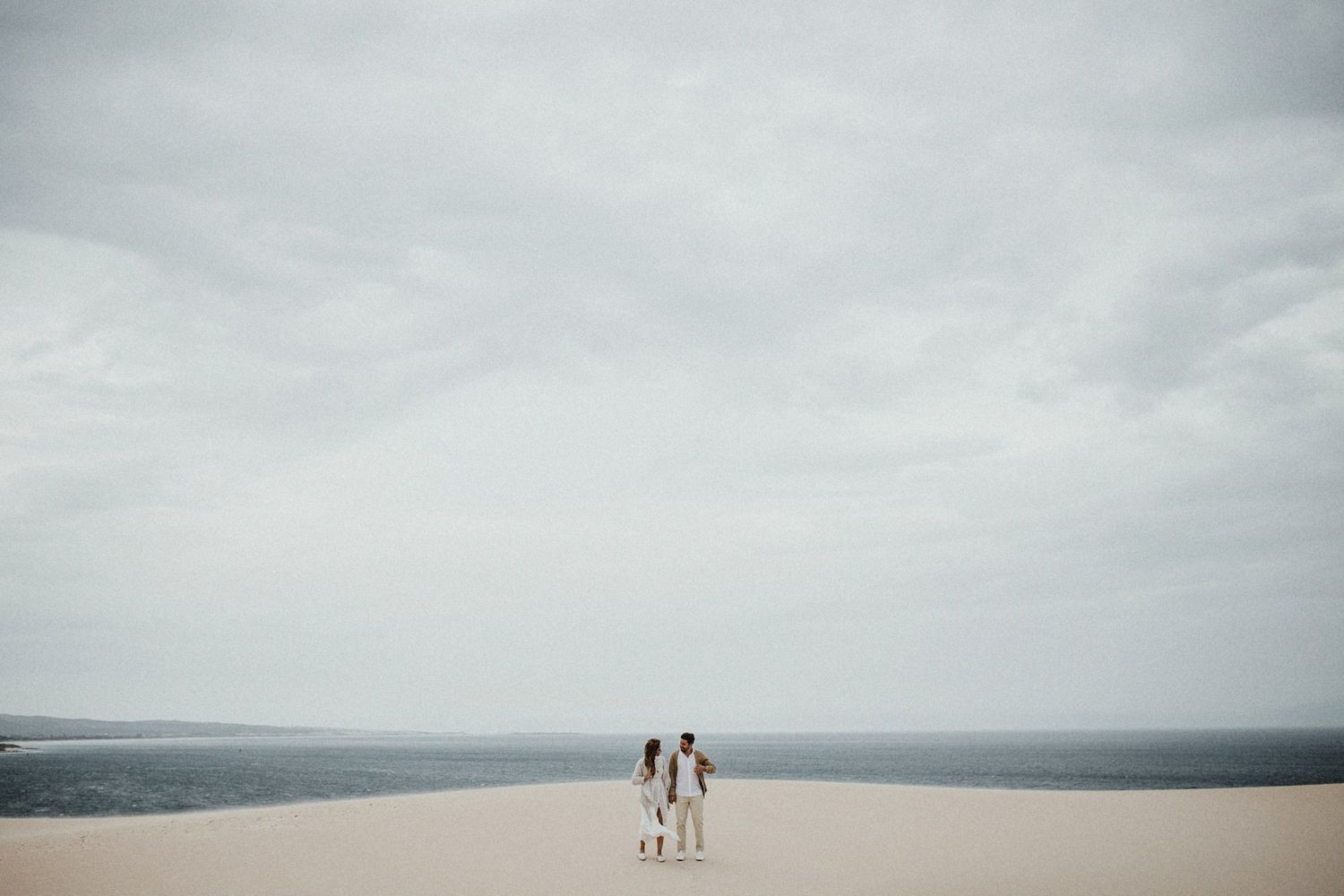 Distant silhouettes of couple standing on sand dunes under overcast sky.