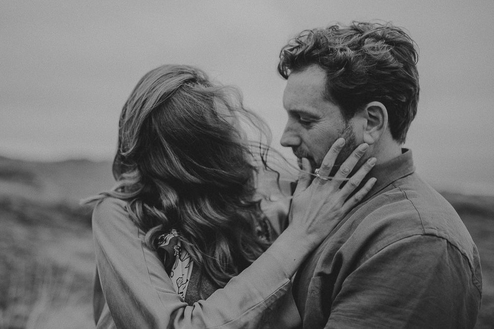 A couple shares a close romantic moment in black and white on a windy hillside.