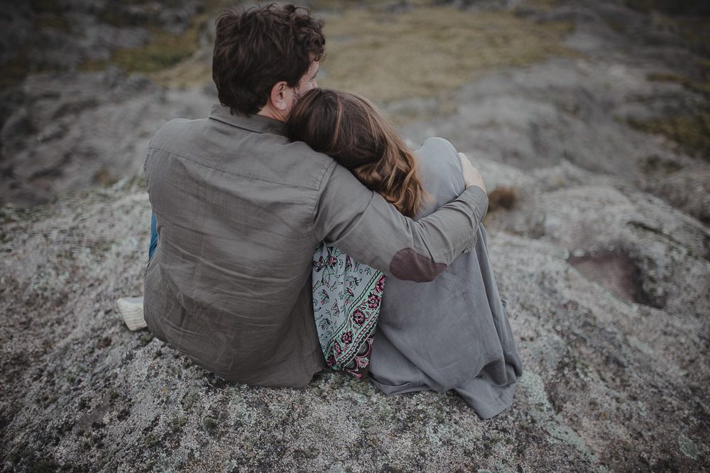 A couple sits close together on rocky terrain at dusk overlooking a scenic landscape.