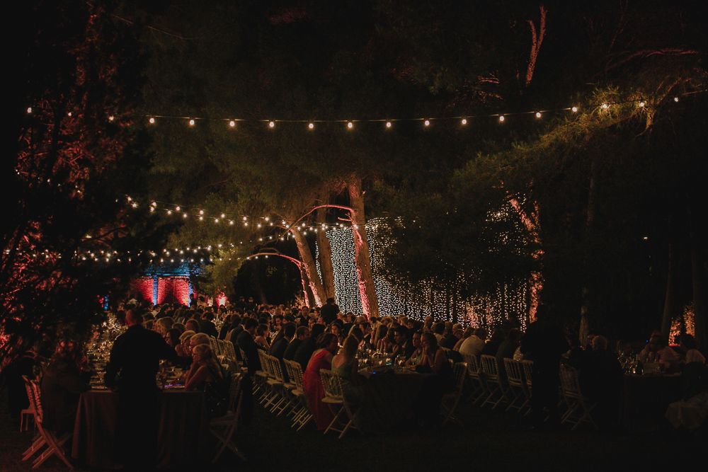 String lights illuminate an outdoor evening wedding reception with a large crowd gathered under trees.