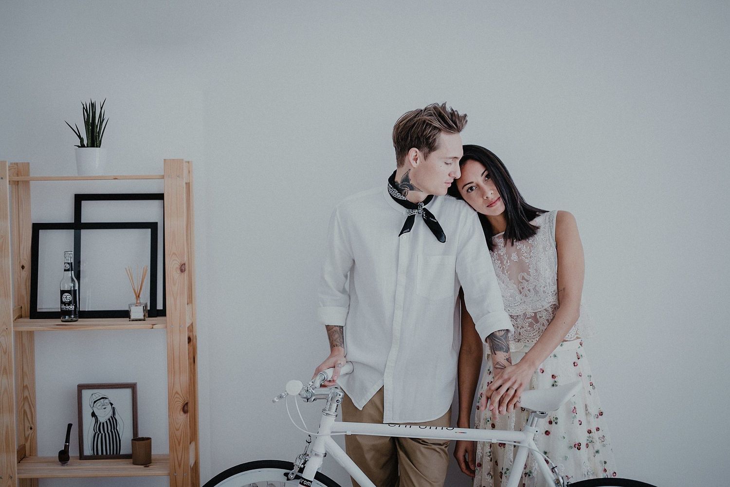 A couple in white outfits standing beside a vintage bicycle in a minimalist room.