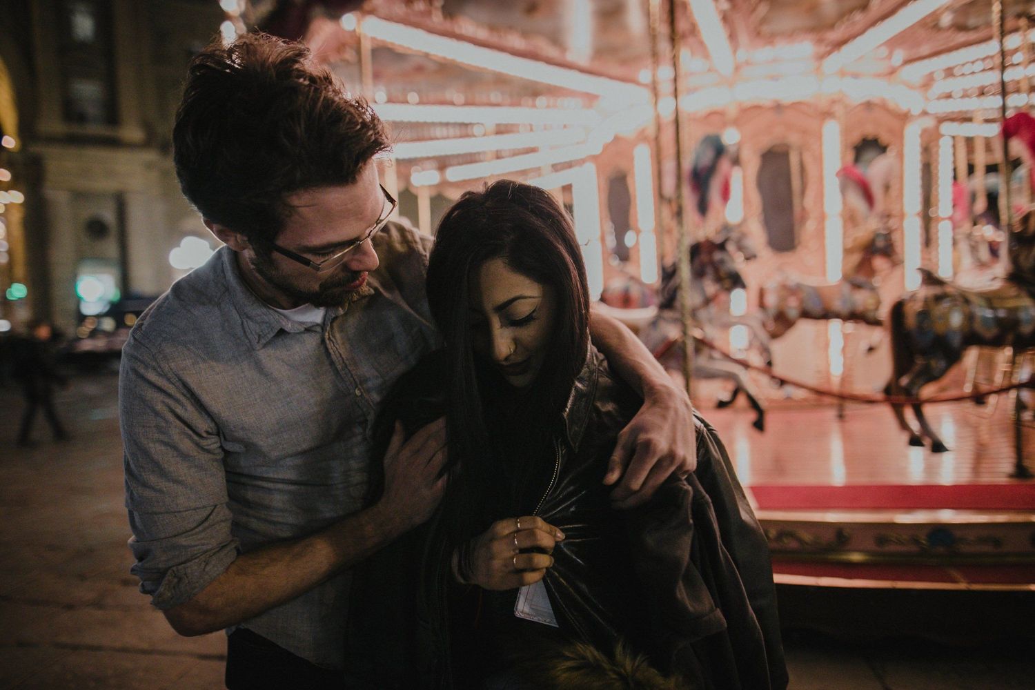 A couple sharing a sweet moment in front of a brightly lit carnival carousel.