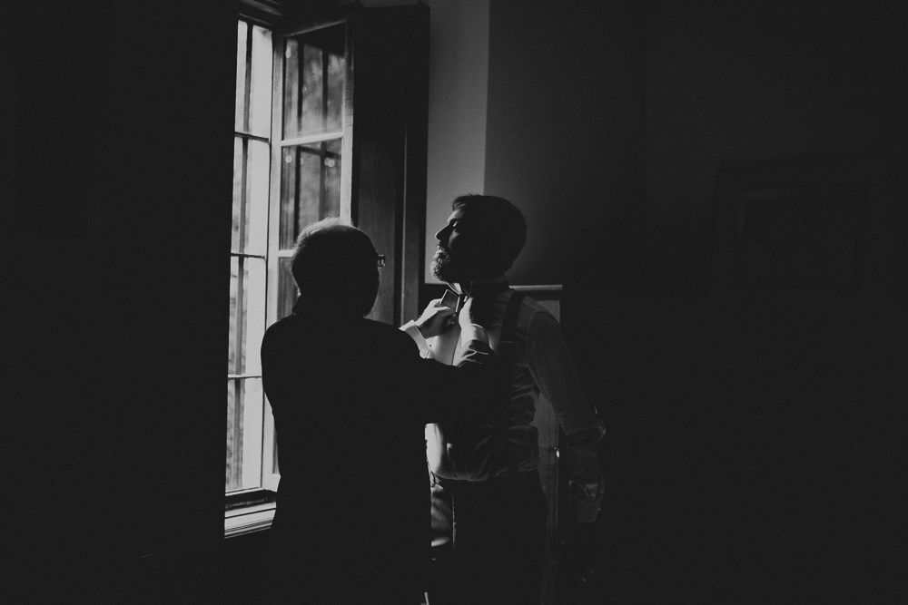 Two silhouettes stand by a window preparing formal attire.