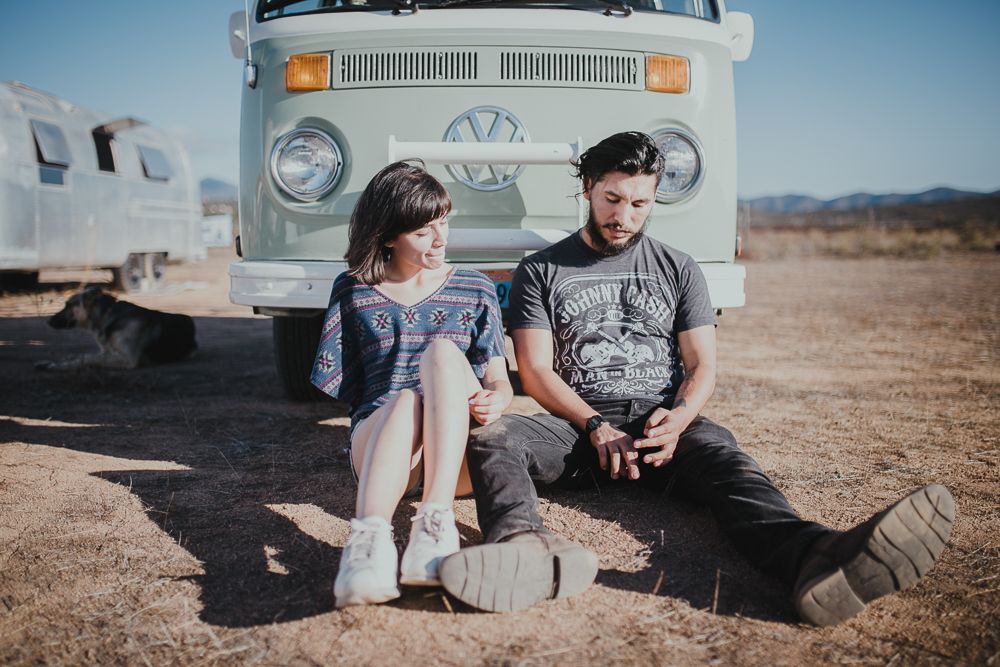 A couple sits together on the ground in front of a vintage Volkswagen van in a desert setting.