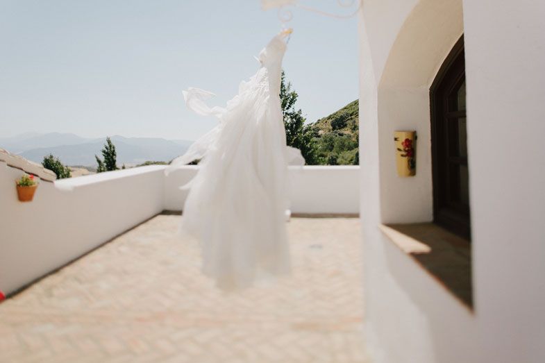 White wedding dress hanging outdoors against white Mediterranean-style architecture with mountains visible.