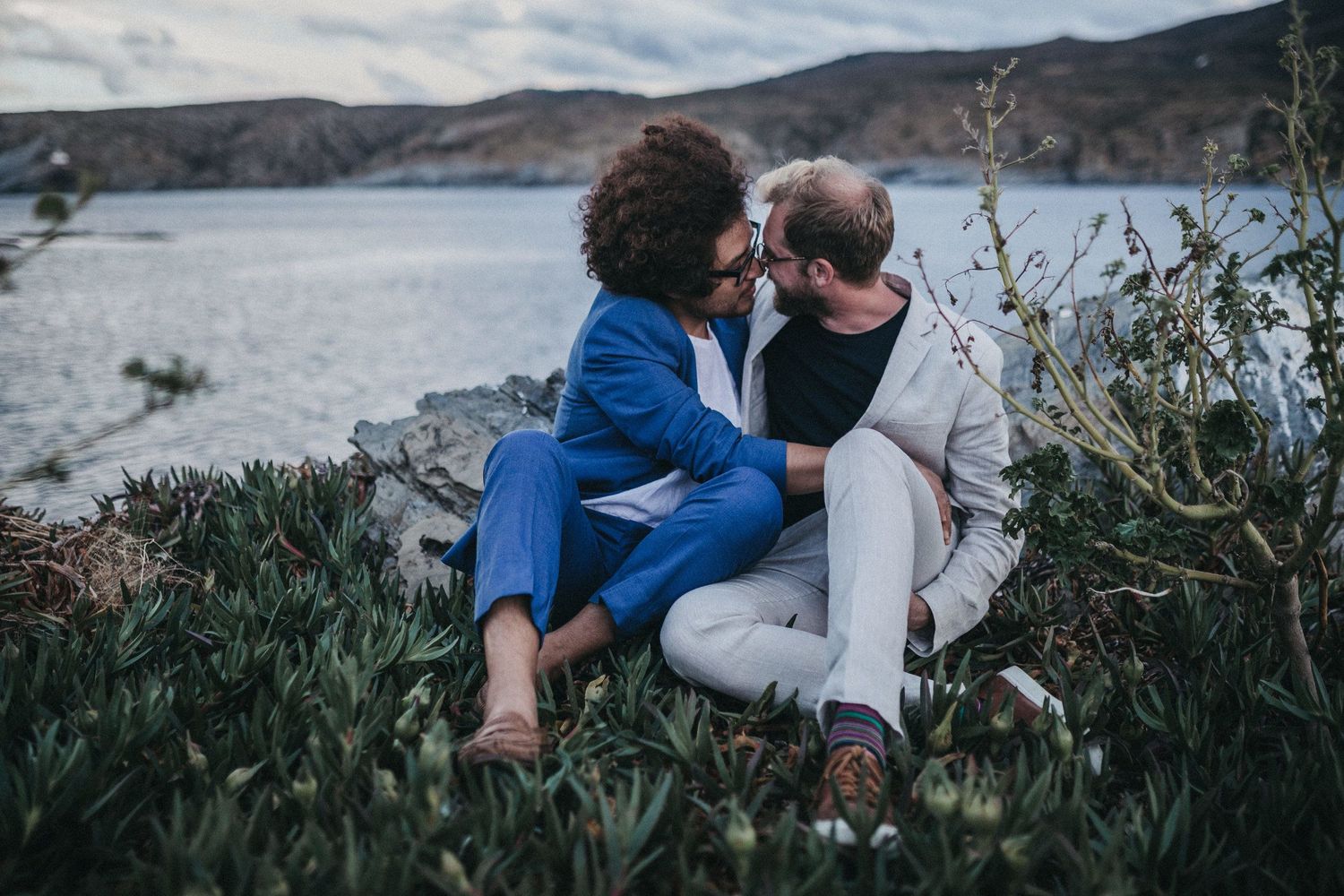 A couple in wedding attire shares an intimate moment by a lake with mountains in the background.