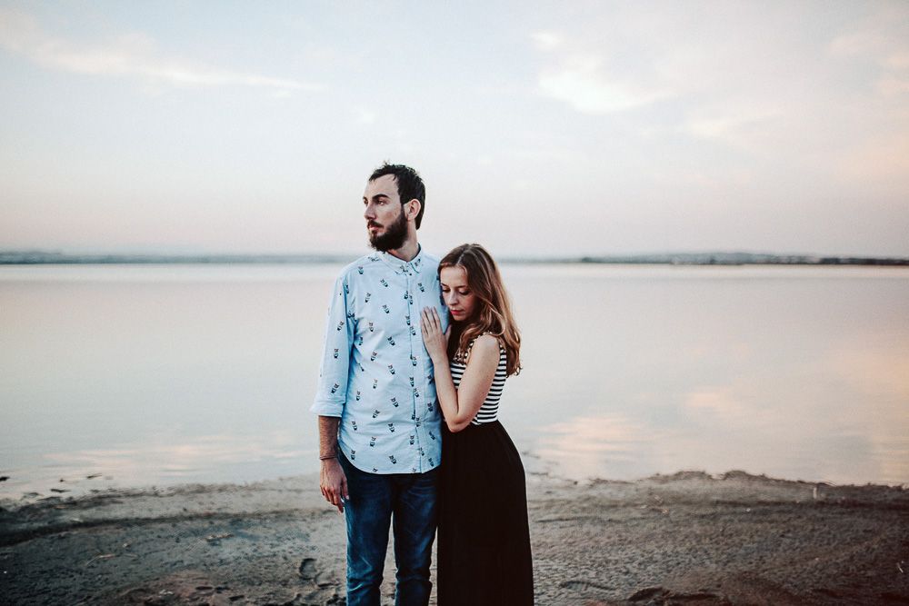 Couple in casual attire standing together on shoreline at sunset.