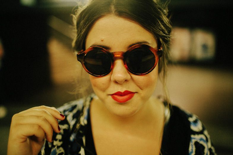 Close-up portrait showing round vintage sunglasses and bright red lipstick against dark background.