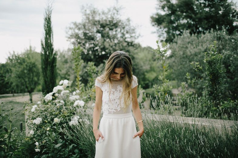 A white lace wedding dress in a garden setting with blooming flowers and cypress trees.