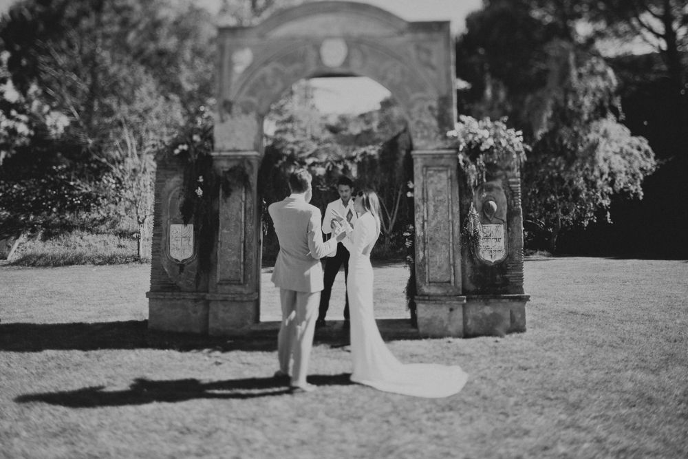 A black and white photo of a couple during their wedding ceremony under a stone archway.