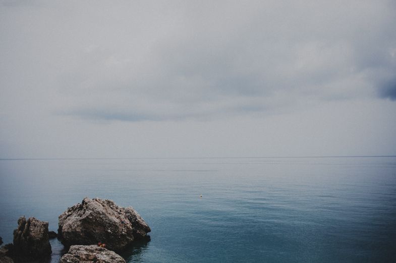 A rocky coastal landscape with calm blue ocean waters under an overcast sky.
