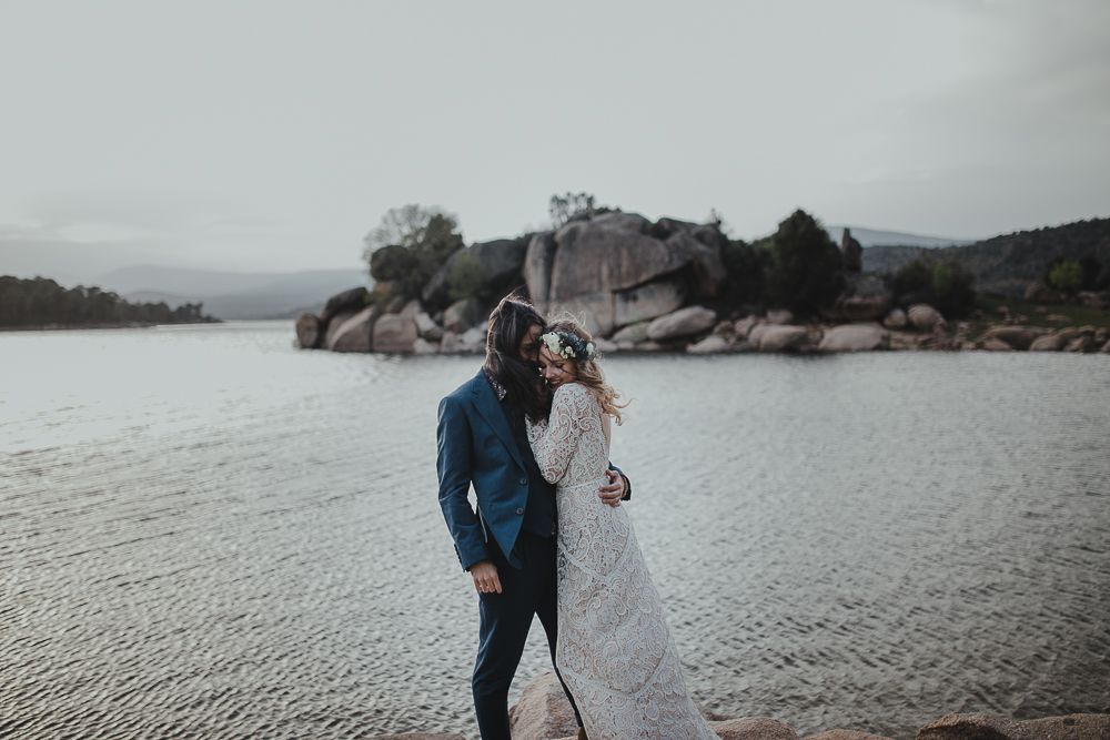 Newlyweds share intimate moment by serene lake surrounded by rocky landscape.