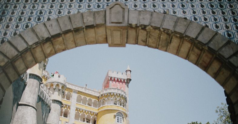 View of Pena Palace towers through a stone archway against bright blue sky.