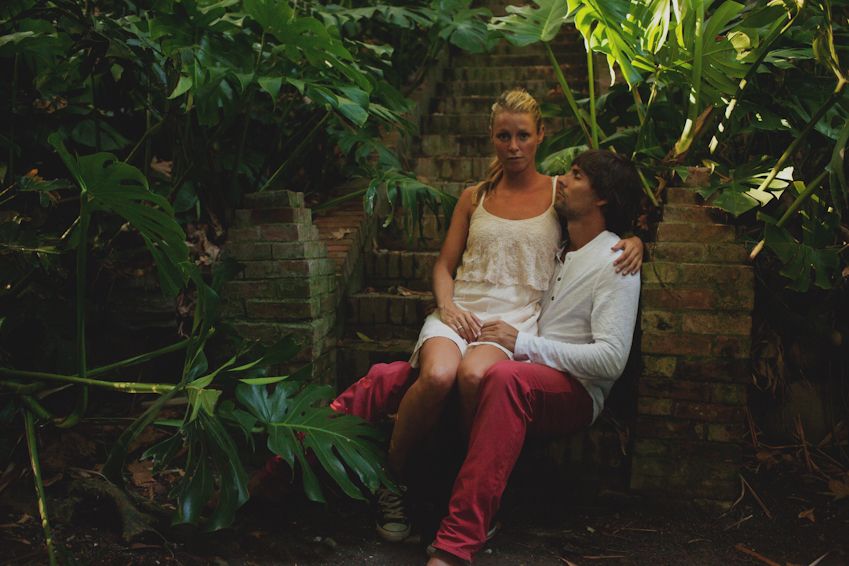 A couple sits casually together on a garden bench surrounded by lush greenery.