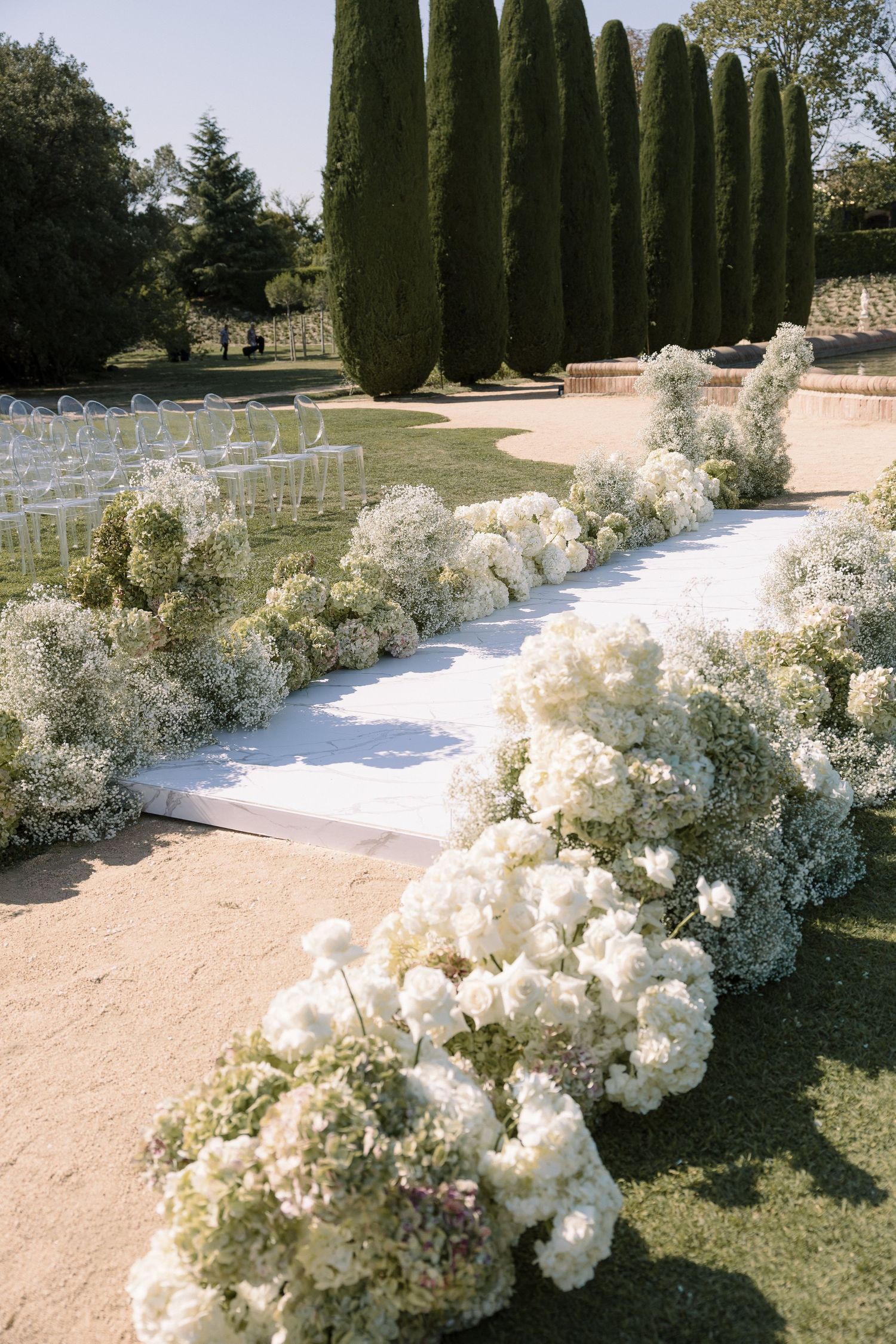 A wedding ceremony aisle lined with lush white flowers and cypress trees.