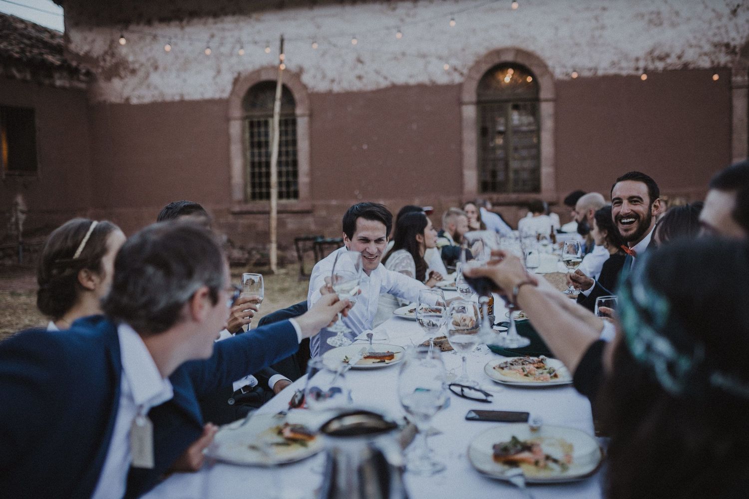 Guests raise glasses in celebration at an outdoor dinner party with rustic decor and warm lighting.