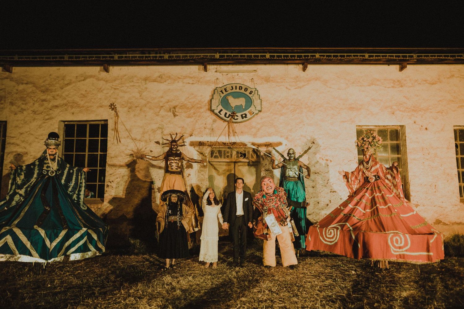 A group in ornate historical costumes poses together against an old stone building facade at night.